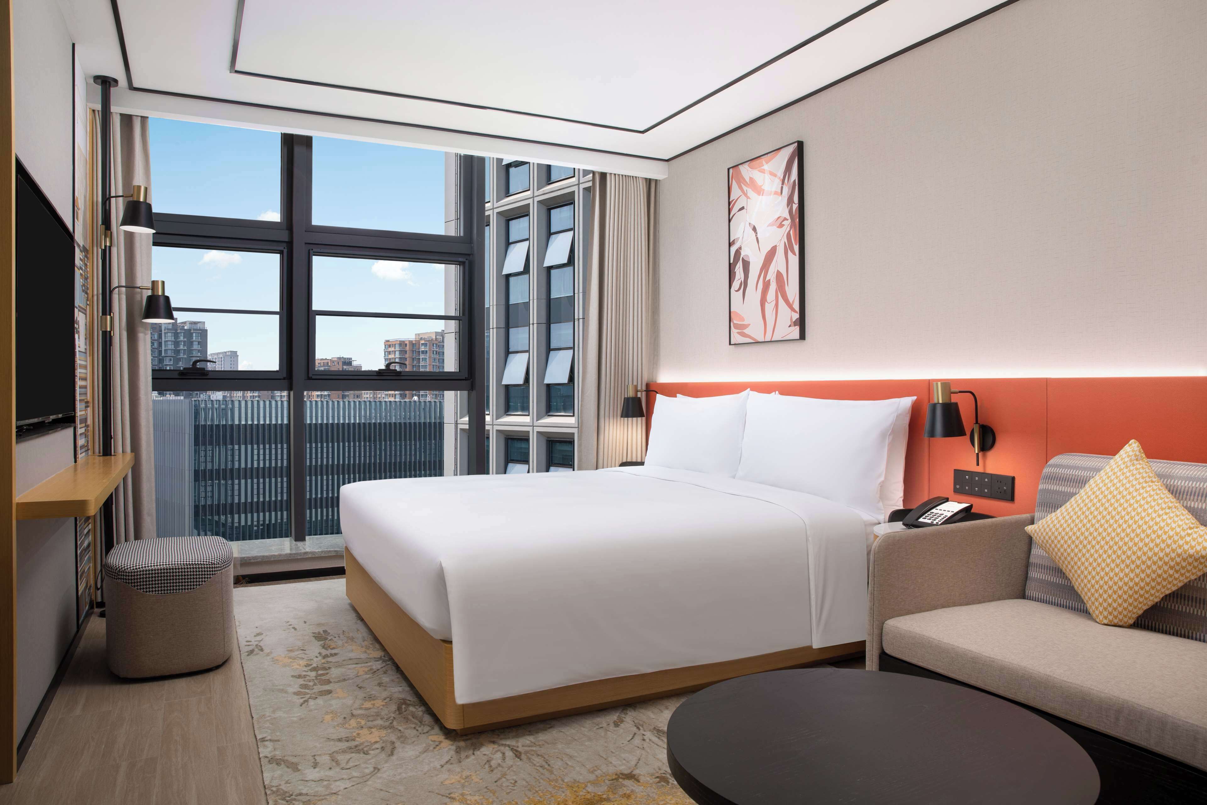 Ein modernes Hotelzimmer mit einem großen Bett, einer Couch, einem kleinen Tisch und Blick auf die Stadt durch große Fenster.