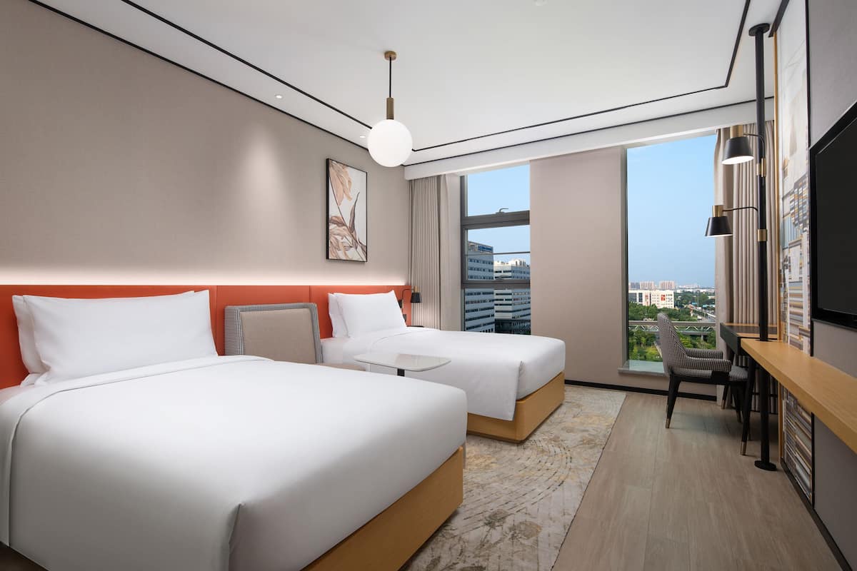 Une chambre d’hôtel avec deux lits, un bureau, une chaise, une télévision et une grande fenêtre donnant sur la ville.