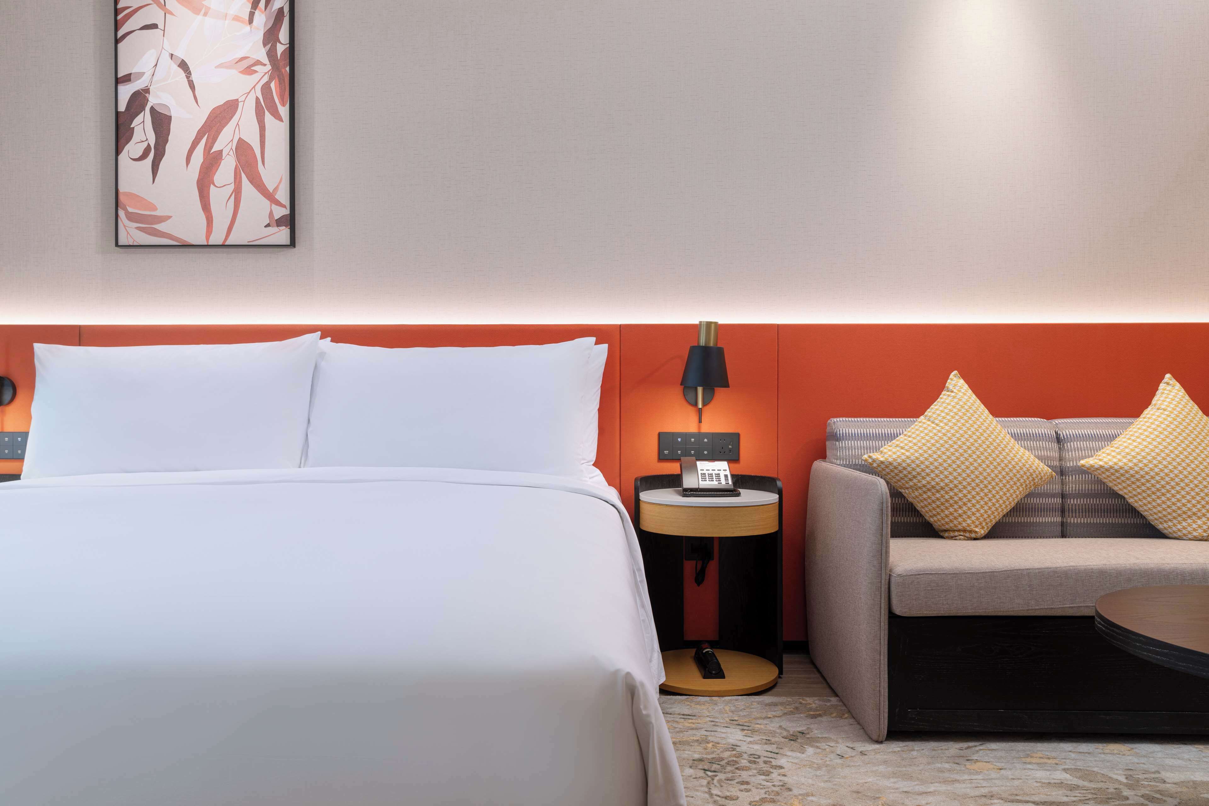 A hotel room with a bed, a sofa, a nightstand with a lamp, and a wall-mounted telephone.