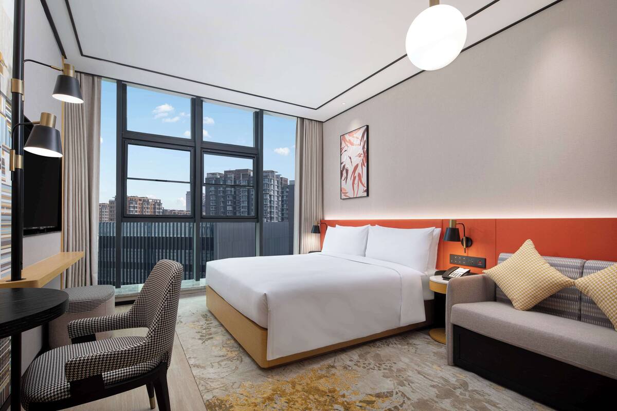 Une chambre d’hôtel moderne, dotée d’un grand lit, d’un canapé, d’un bureau et d’une chaise. Une grande fenêtre offre une vue sur la ville.