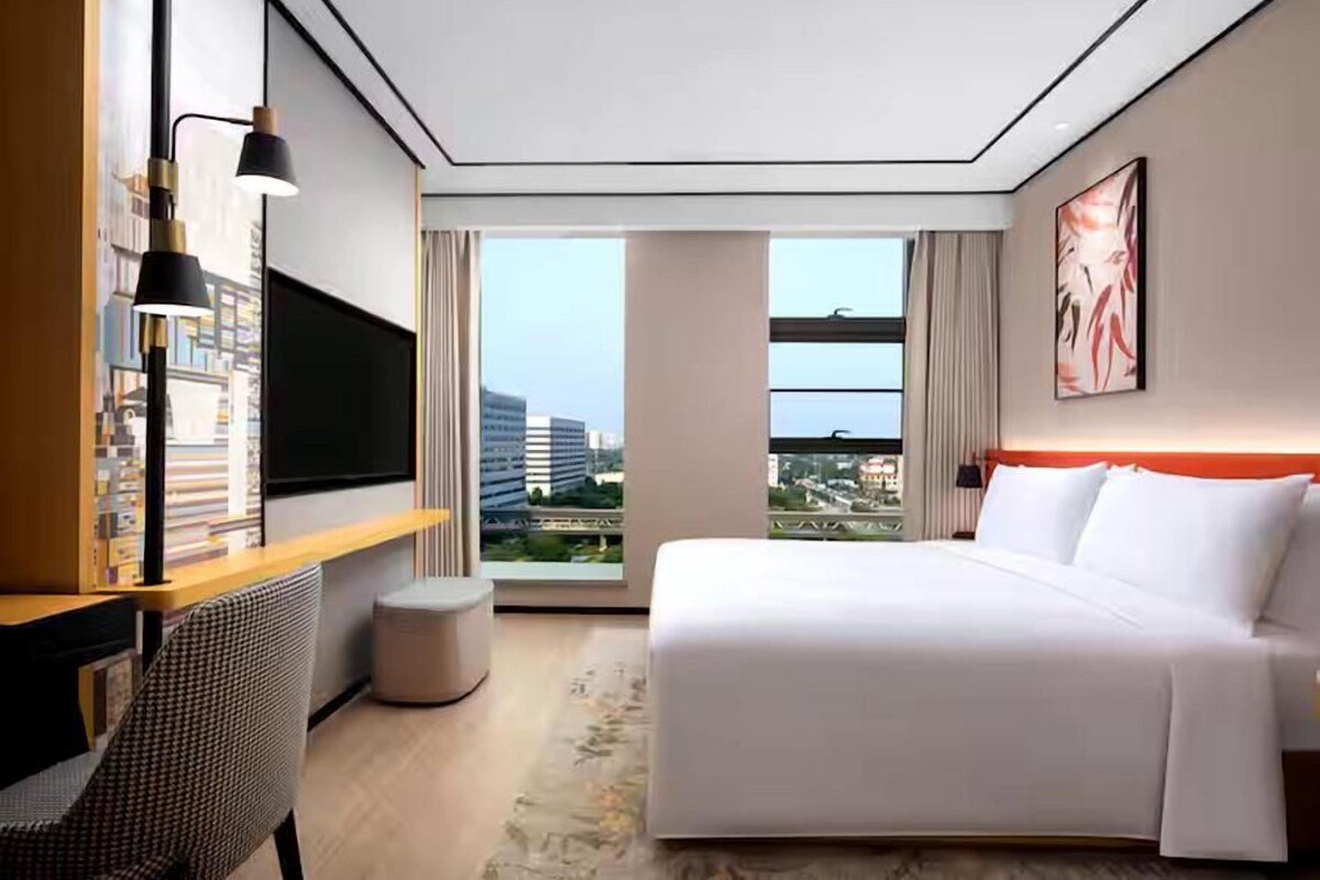 A hotel room with a large bed, a desk with a chair, a TV, and a view of the city through large windows.