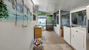 A compact kitchen with a washing machine, a wooden cabinet, and a basket of laundry. - Relaxing Retreat 2BR Oasis w/ Pool and Outdoor BBQ (Pembroke Pines)