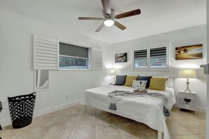 Ein Schlafzimmer mit einem Bett, zwei Nachttischen, einem Deckenventilator und zwei Fenstern mit Fensterläden.