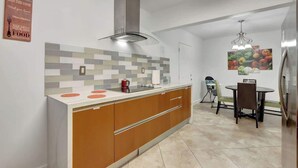 A modern kitchen with a central island, a built-in oven, and a stainless steel range hood. There is a dining area with a table and chairs, and a wall-mounted artwork above the table. - Relaxing Retreat 2BR Oasis w/ Pool and Outdoor BBQ (Pembroke Pines)