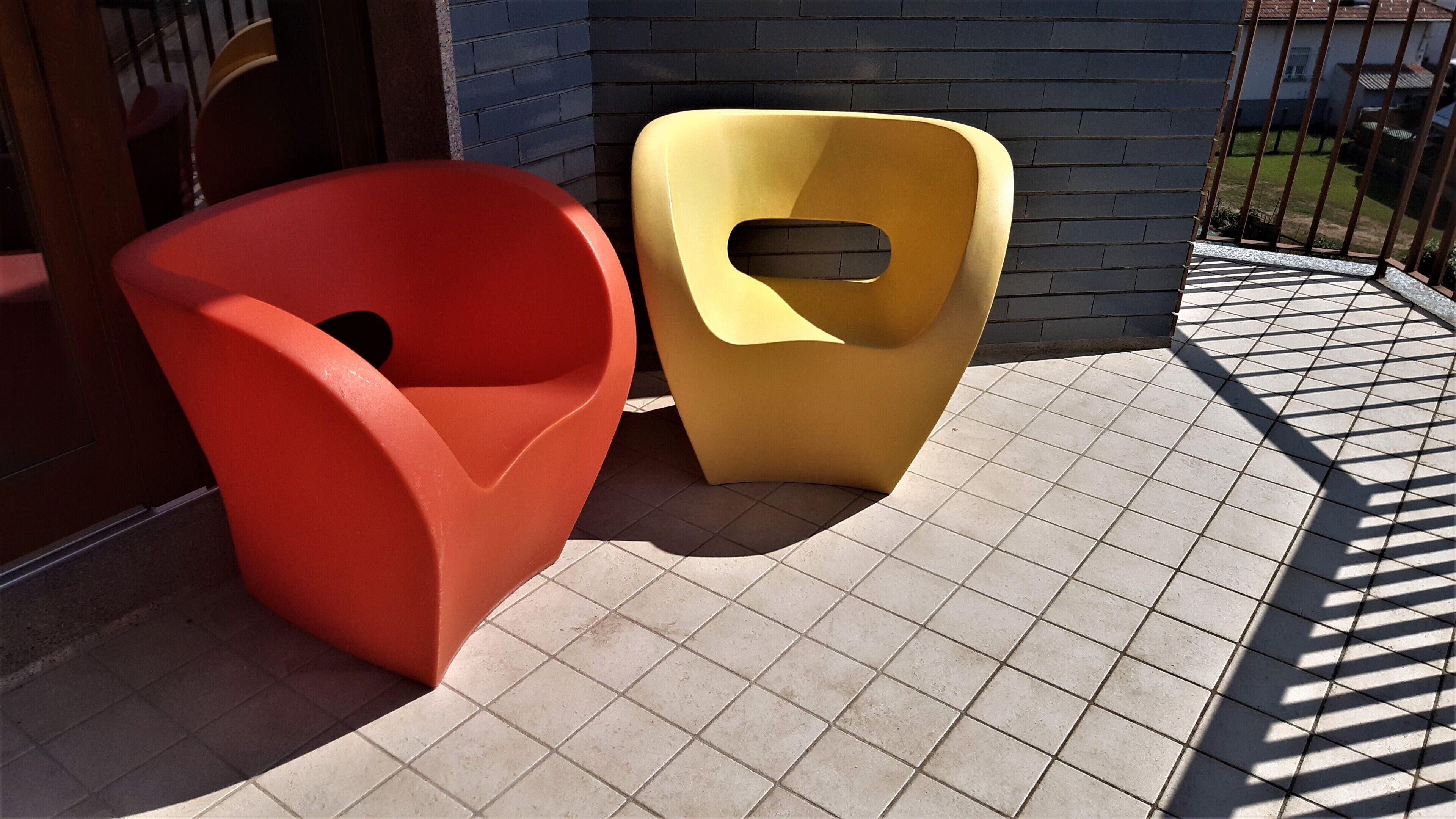 Due moderne sedie dallo stile scultoreo su un patio piastrellato, con un muro in mattoni e un recinto metallico sullo sfondo.