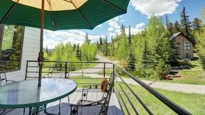 A balcony with a glass table and chairs, an umbrella, and a view of a forested area with a house in the background. - Beautiful Views, Awesome Amenities, 195 Eastridge (Dillon)