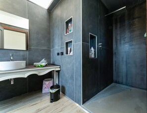 Ein modernes Badezimmer mit einem großen Spiegel, einem weißen Waschbecken, einem Waschtisch, einer Dusche mit dunkler Tür und zwei kleinen Ablagen mit Pflegeprodukten.