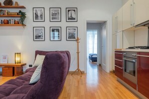 A modern living room with a purple sofa, a wooden side table, and a floor lamp. There are framed pictures on the wall and a coat rack. A kitchen with wooden cabinets and a stove is visible in the background. - La Corte by Wonderful Italy (Turin)