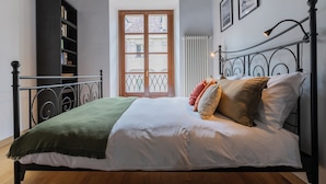 Una cama bien hecha con una manta verde, un edredón blanco y varias almohadas. El suelo es de madera, el cabecero de metal negro, y hay una ventana con vistas a edificios.