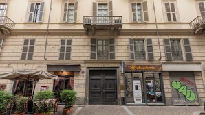 Un edificio con entrada central, un pequeño espacio al aire libre con mesas y sillas, y un escaparate con un letrero.