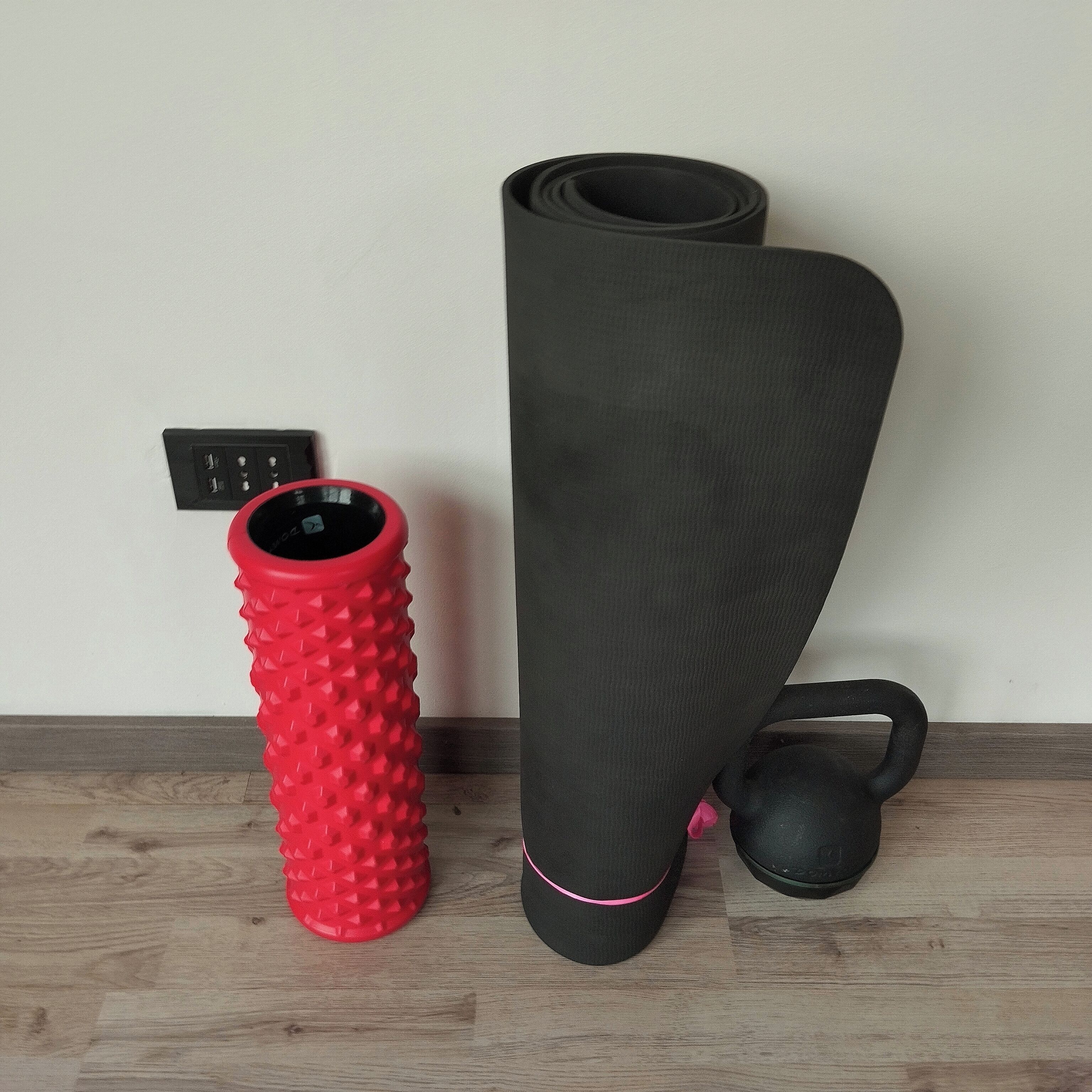 Un oggetto rosso con texture, un oggetto nero arrotolato e un oggetto nero con un cinturino rosa su un pavimento di legno, con una parete bianca sullo sfondo.