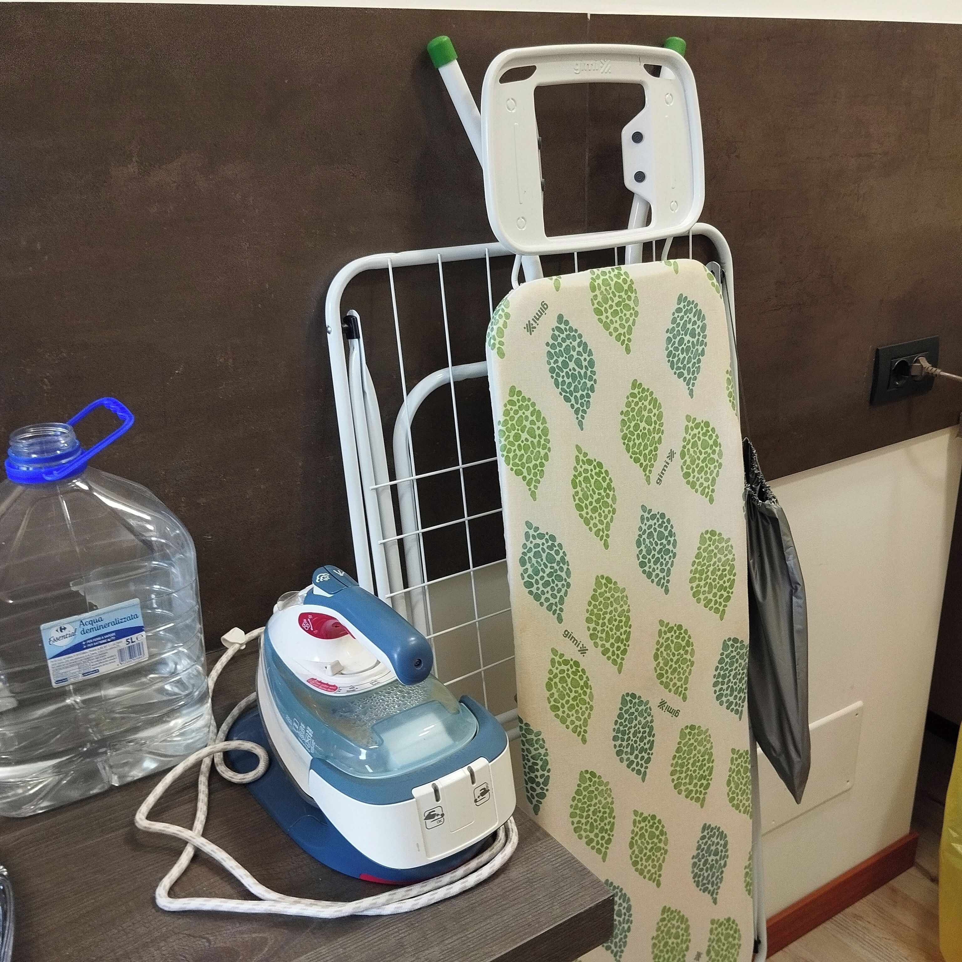 Una tabla de planchar con diseño de hojas verdes, un planchador y una botella de agua sobre una superficie de madera.