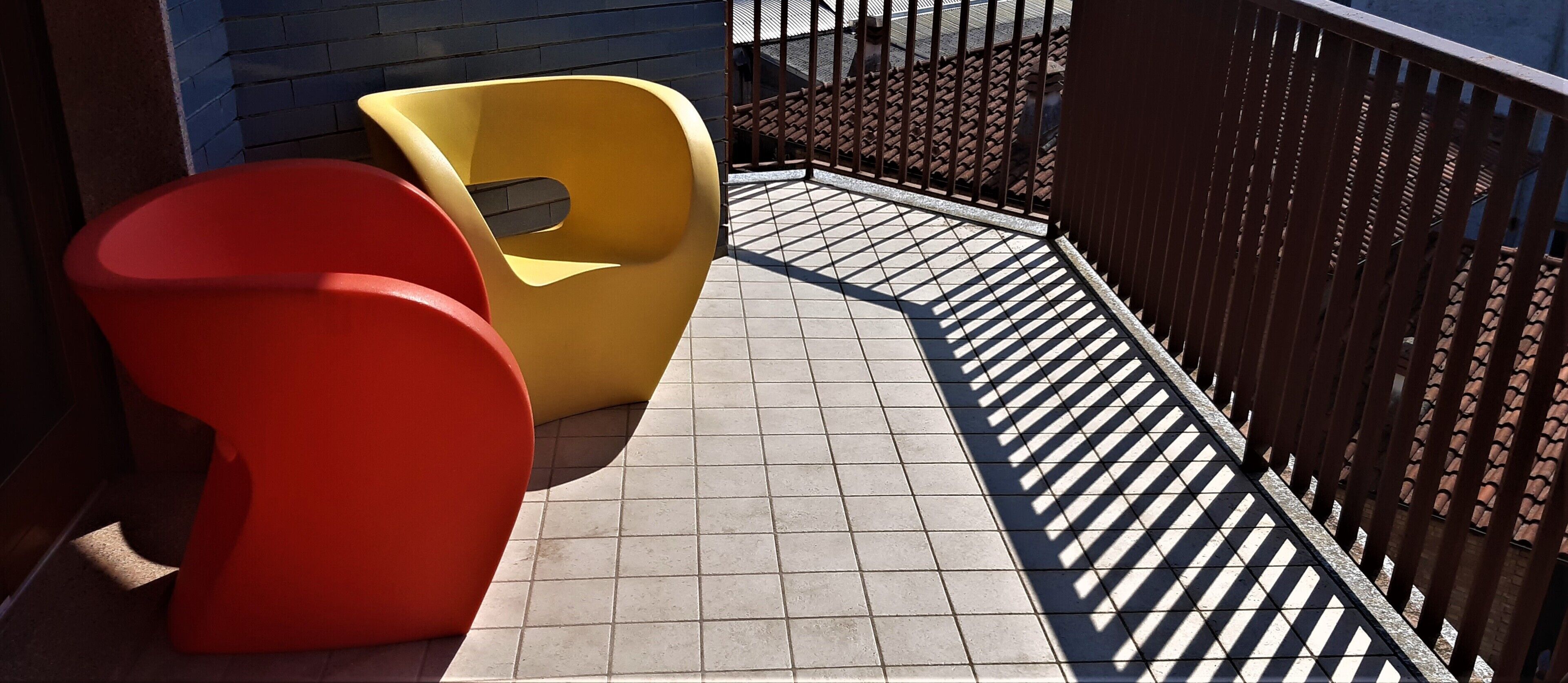 A tiled balcony with two chairs, one red and one yellow, and a metal railing.