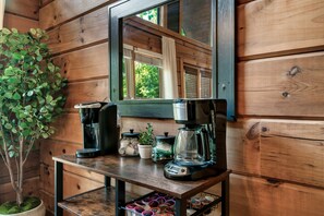 Interior de cabana de madeira, com um cantinho de café, uma planta em vaso e uma janela com vista para o verde.
