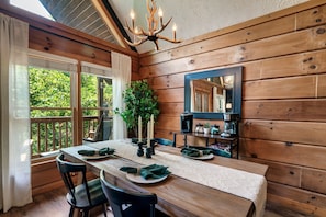 A wooden dining room with a table set for four, a chandelier, and a view of greenery outside.
