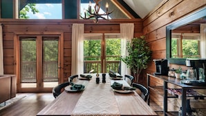 Una cabaña de madera con una mesa de comedor preparada para cuatro personas, una araña y una zona de cocina.