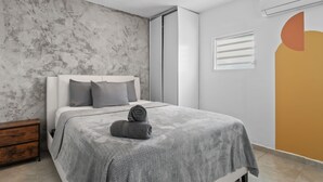 Un dormitorio moderno con cama, mesita de noche de madera, pared de hormigón y un amplio ventanal con persianas.