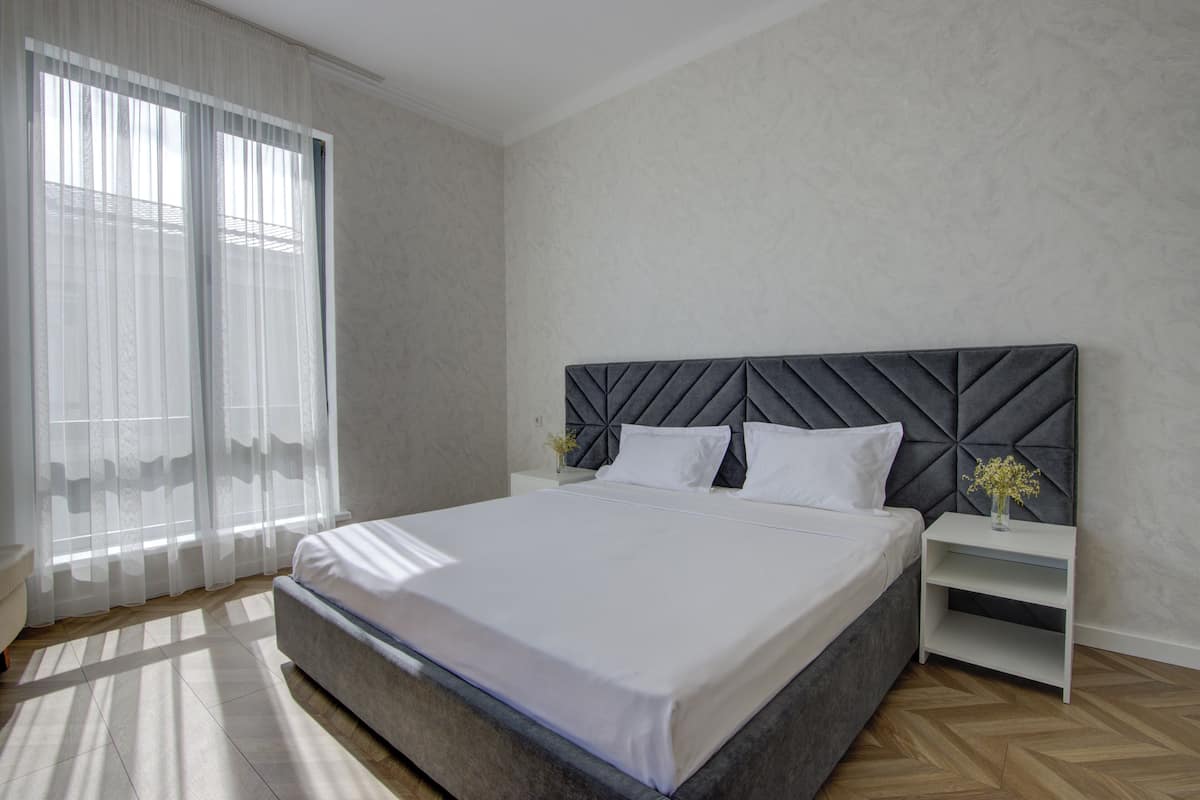 A bedroom with a bed, a nightstand, a vase with flowers, and sheer curtains.