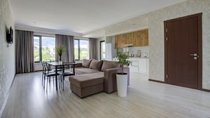 Een moderne woonkamer met een hoekbank, een eettafel en stoelen. Er is een groot raam met gordijnen, een potplant en een keuken in de achtergrond.