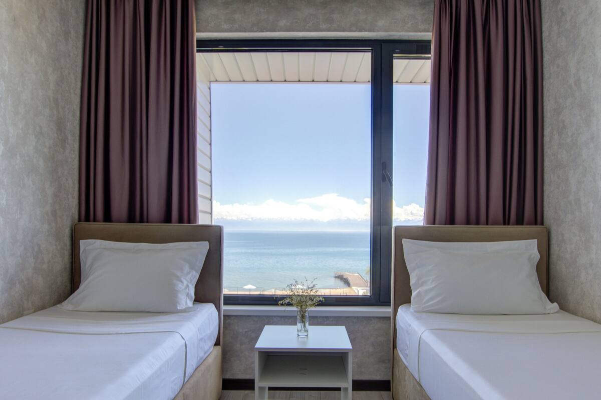 A hotel room with two beds, a small table, and a window with a view of the sea.