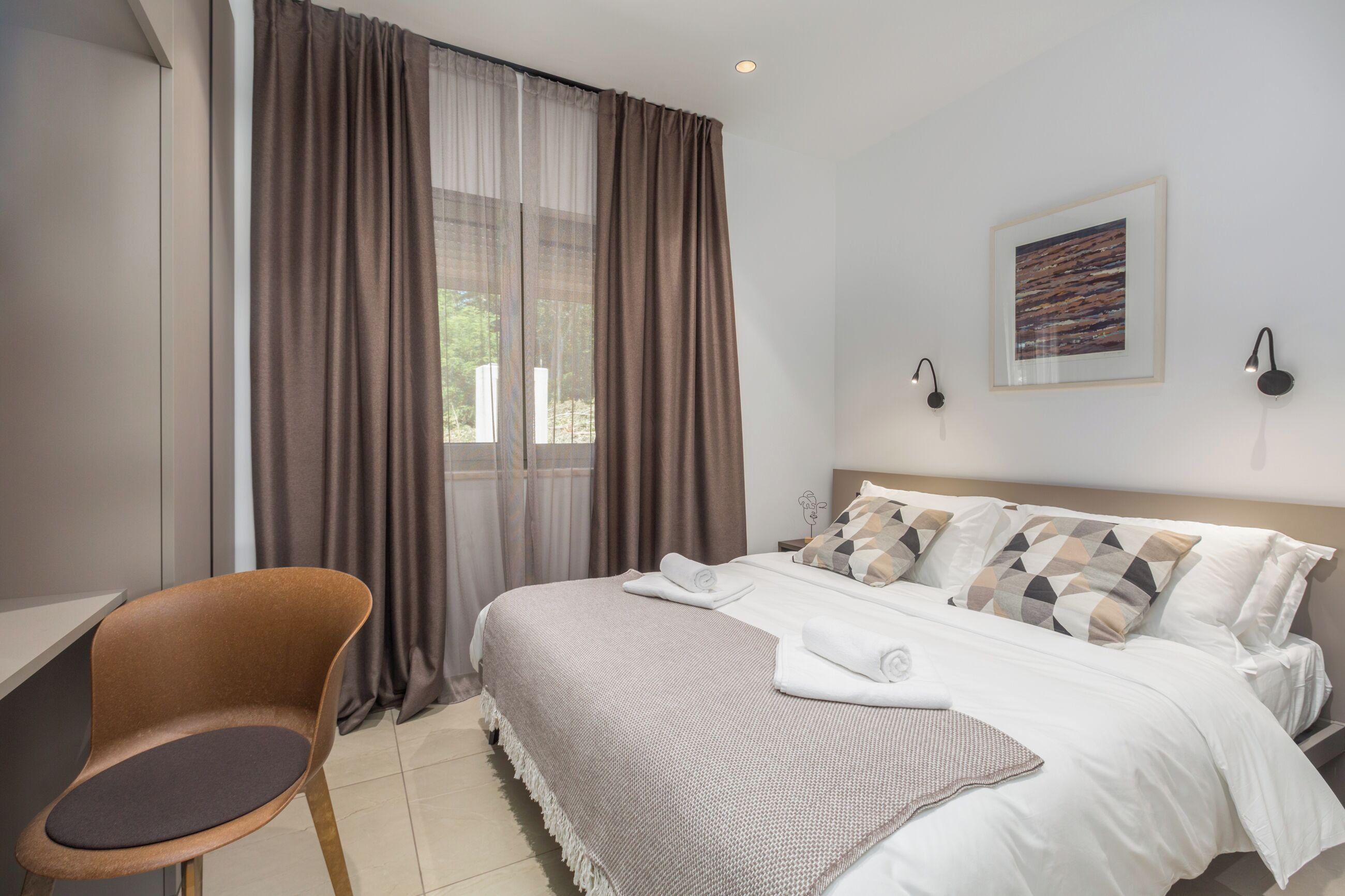 A hotel room with a bed, a chair, a window with curtains, and a framed picture on the wall.