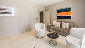 Een moderne woonkamer met een bank, een witte fauteuil en een salontafel. Aan de muren hangen abstracte schilderijen en er is een deur naar een andere kamer.
