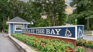 A sign for 'Angola by the Bay' with a sailboat graphic, surrounded by a brick wall and red flowers.