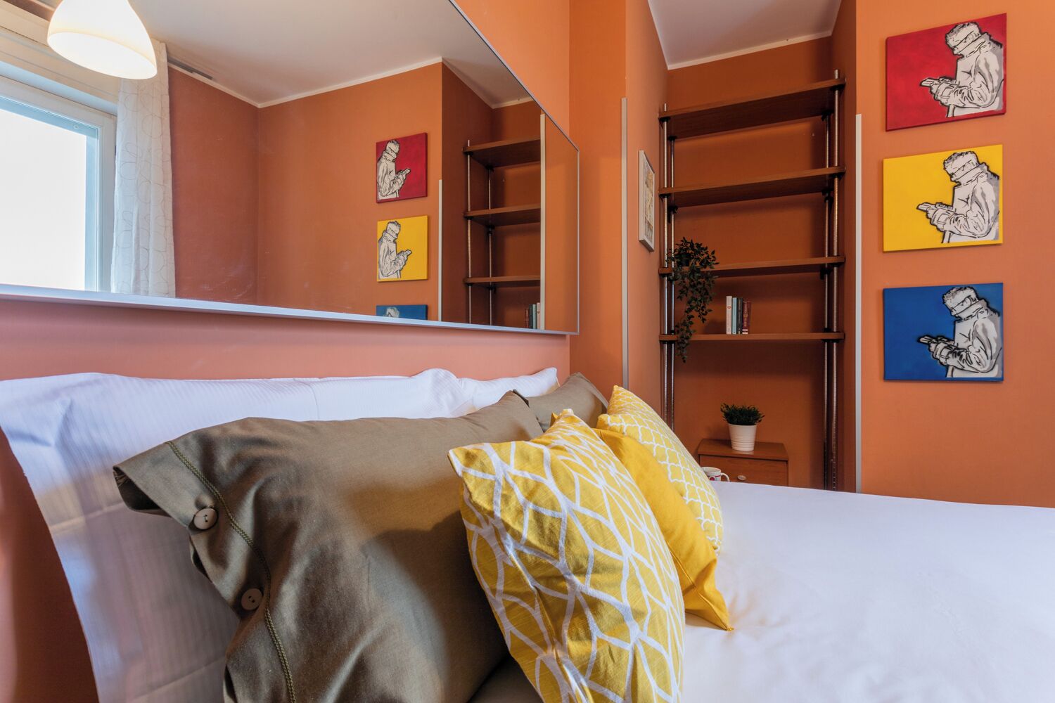 Un dormitorio con cama, almohadas, una mesita de noche, un espejo y una estantería con libros y obras de arte.