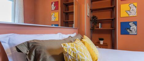 Un dormitorio con cama, almohadas, una mesita de noche, un espejo y una estantería con libros y obras de arte.