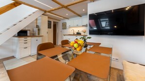 A modern kitchen with white cabinets, a dining area with orange chairs, and a large flat-screen TV mounted on the wall.