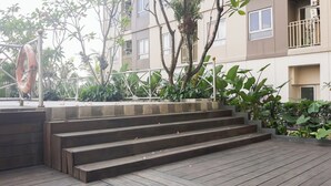 A set of wooden steps leading up to a concrete platform with a metal railing, surrounded by greenery and a building with multiple windows and balconies. - Comfortable And Tidy Studio At Titanium Square Apartment (Jakarta)