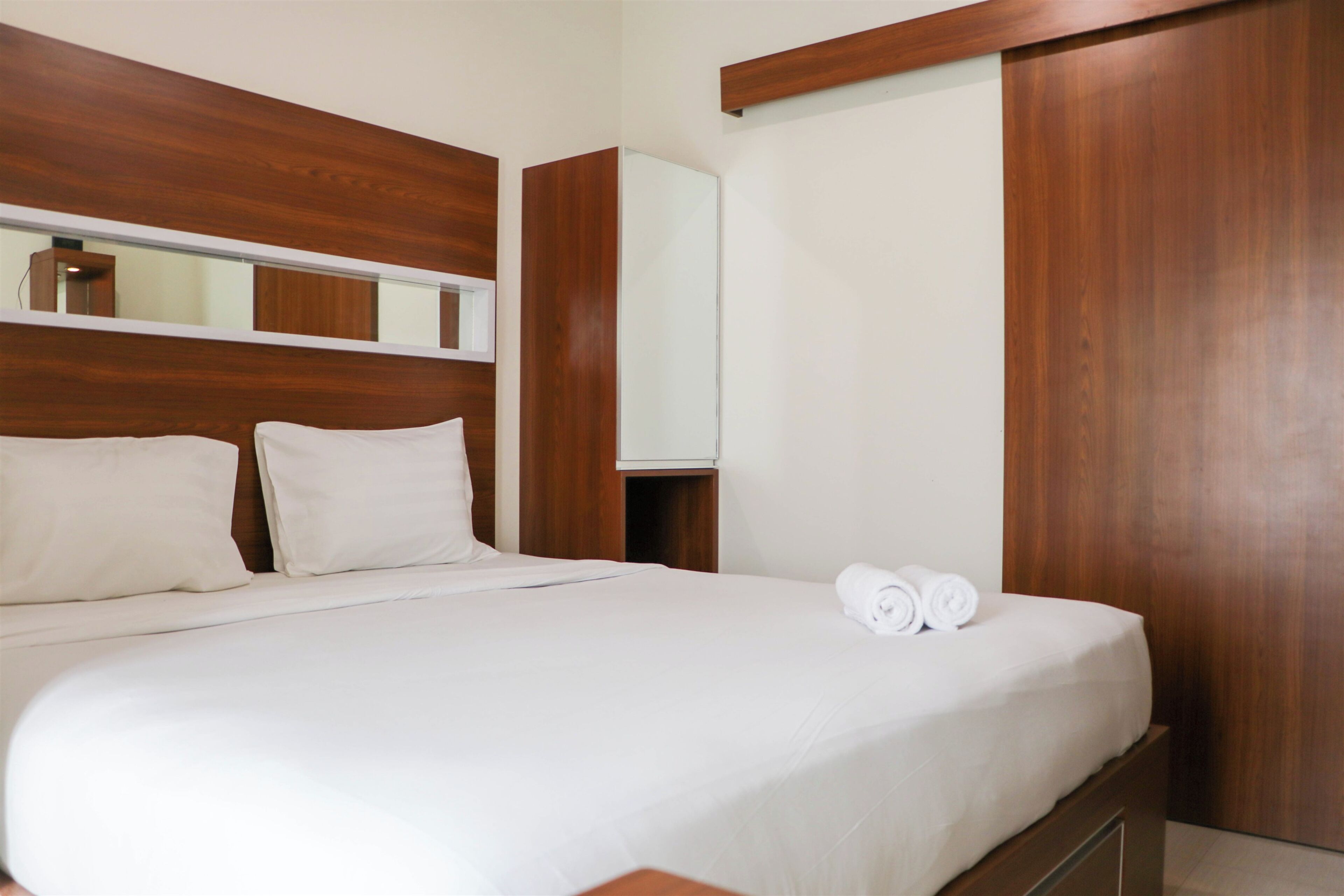 A neatly made bed with white linens and pillows, a wooden headboard, a mirror, and a wooden wardrobe.