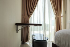 A hotel room with a bed, a small round table, a mounted shelf, and sheer curtains. - Restful 1Br Without Living Room Grand Kamala Lagoon Apartment (Bekasi)