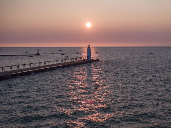 A pier extending into calm waters with a lighthouse at the end, boats in the distance, and a clear sky.
