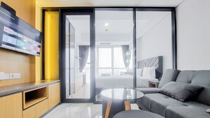 A modern hotel room with a flat-screen TV displaying various streaming services, a sofa, a glass coffee table, and a view of the adjacent bedroom through a sliding door. - Good And Restful 1Br At The Smith Alam Sutera Apartment (Tangerang)