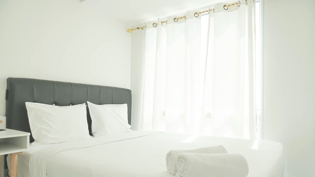A neatly made bed with white linens and a rolled towel, a bedside table, and a window with sheer curtains. - Restful And Comfy 2Br Tokyo Riverside Pik 2 Apartment (Teluknaga)