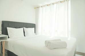 A neatly made bed with white linens and a rolled towel, a bedside table, and a window with sheer curtains. - Restful And Comfy 2Br Tokyo Riverside Pik 2 Apartment (Teluknaga)
