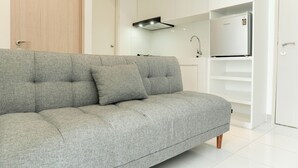 A modern living room with a grey sofa, a built-in kitchen area, and a white door. - Restful And Comfy 2Br Tokyo Riverside Pik 2 Apartment (Teluknaga)