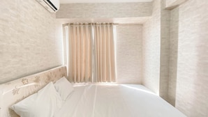 A neatly made bed with white linens, a headboard with floral patterns, and a window with vertical blinds.