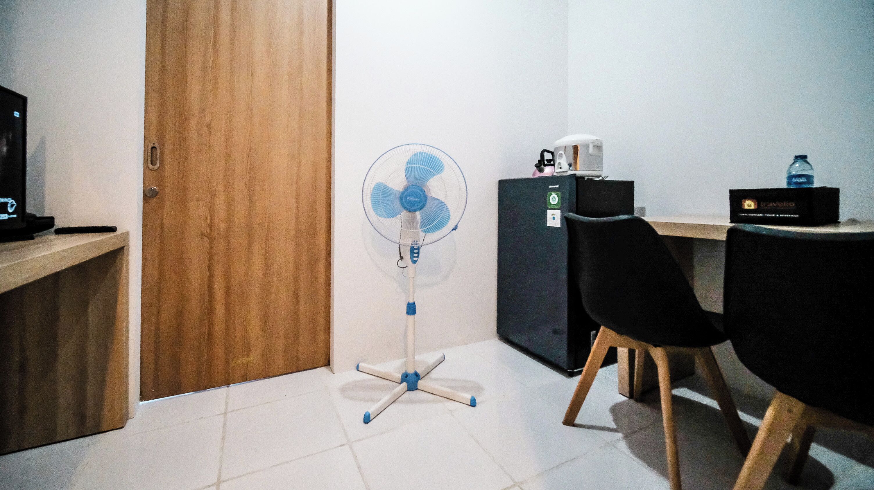 A small room with a wooden door, a standing fan, a desk with a computer, a chair, and a refrigerator.