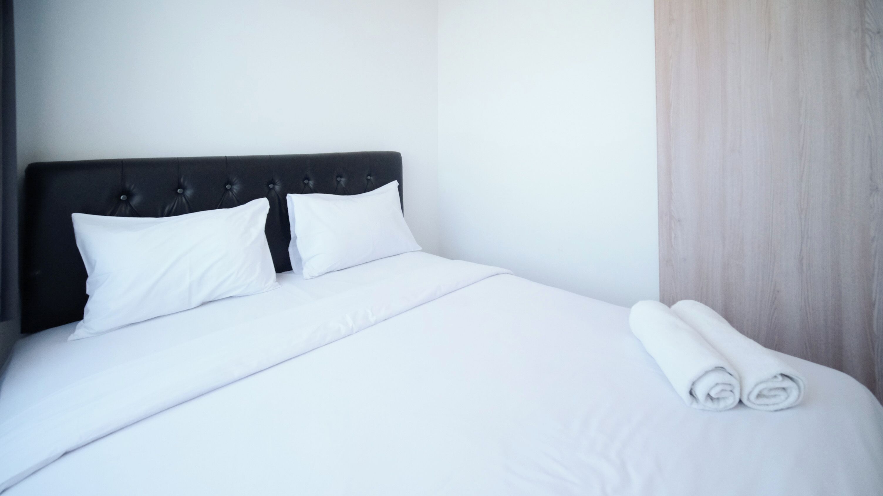 A neatly made bed with white linens and pillows, a dark headboard, and a rolled towel on the side.