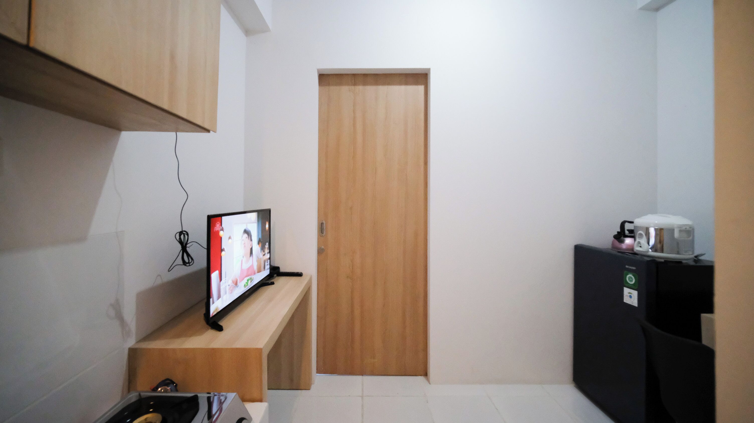 Ein kleiner, schmales Zimmer mit einer Holztür, einem Fernseher auf einem Holzständer und einer Kaffeemaschine auf einem schwarzen Schrank.