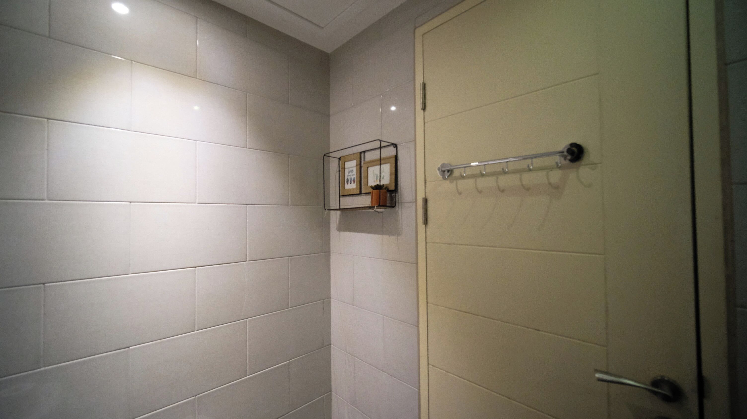 A bathroom with white tiled walls, a door with a towel rack, and a small shelf with framed pictures and a potted plant.
