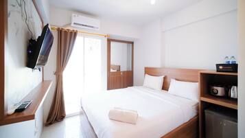 Habitación de hotel con cama, televisor montado en la pared, armario, kitchenette pequeño y ventana con cortinas.