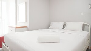 Un letto rifatto con lenzuola bianche e un asciugamano arrotolato, un comodino e una finestra con tende.