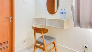 A wooden chair with a grey cushion in a room with a wooden door, a mirror, and a shelf with water bottles. - Good Studio (Free Ipl & Utilities) Grand Palace Kemayoran Apartment (Jakarta)