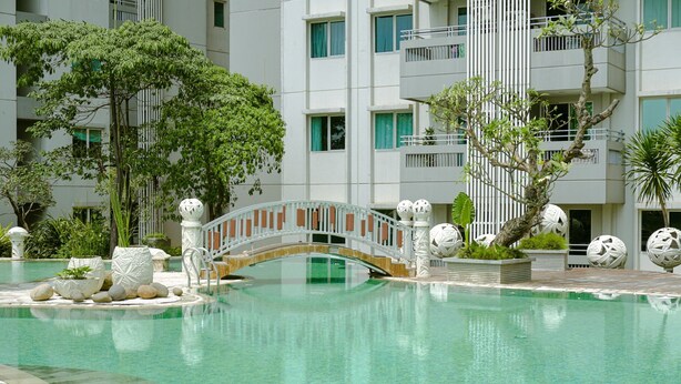 Une piscine avec un pont, entourée de verdure et un bâtiment moderne doté de balcons.