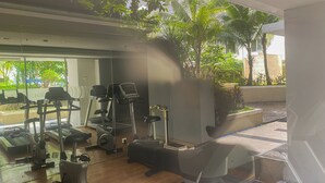 Ein Fitnessstudio mit verschiedenen Trainingsgeräten, darunter Laufbänder und Crosstrainer, mit Blick auf Palmen und Grünflächen im Freien.
