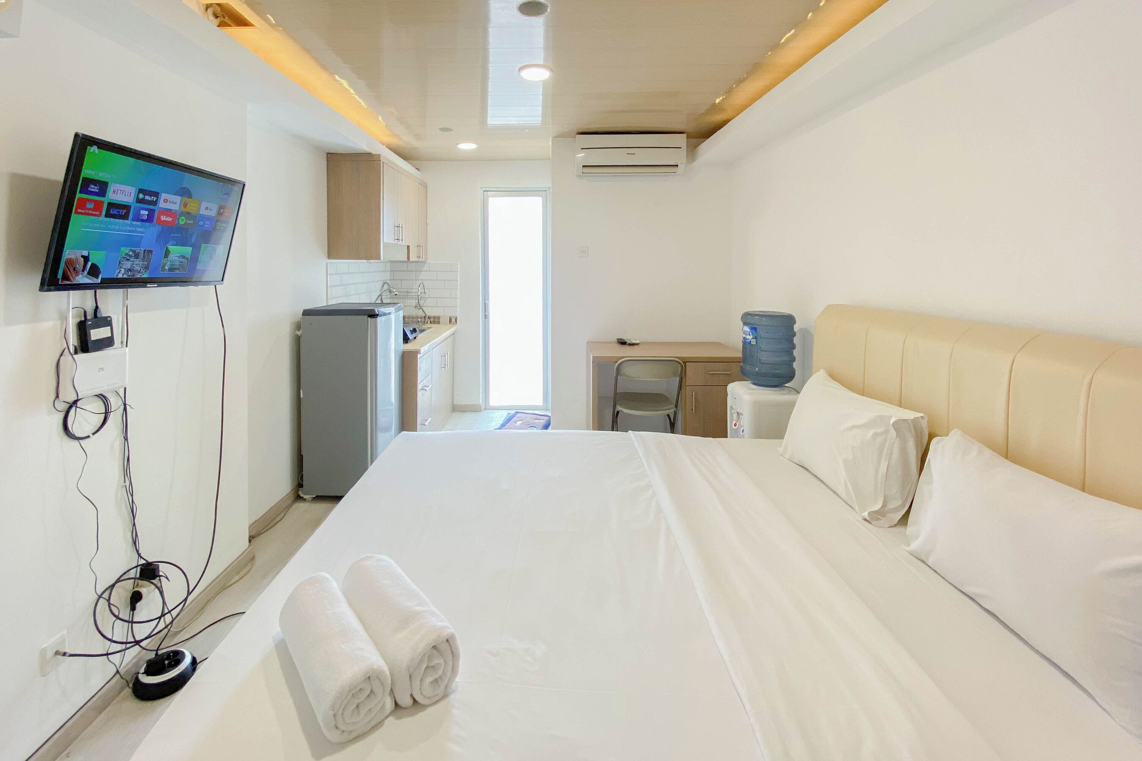 A hotel room with a bed, a TV mounted on the wall, a desk, and a kitchenette.