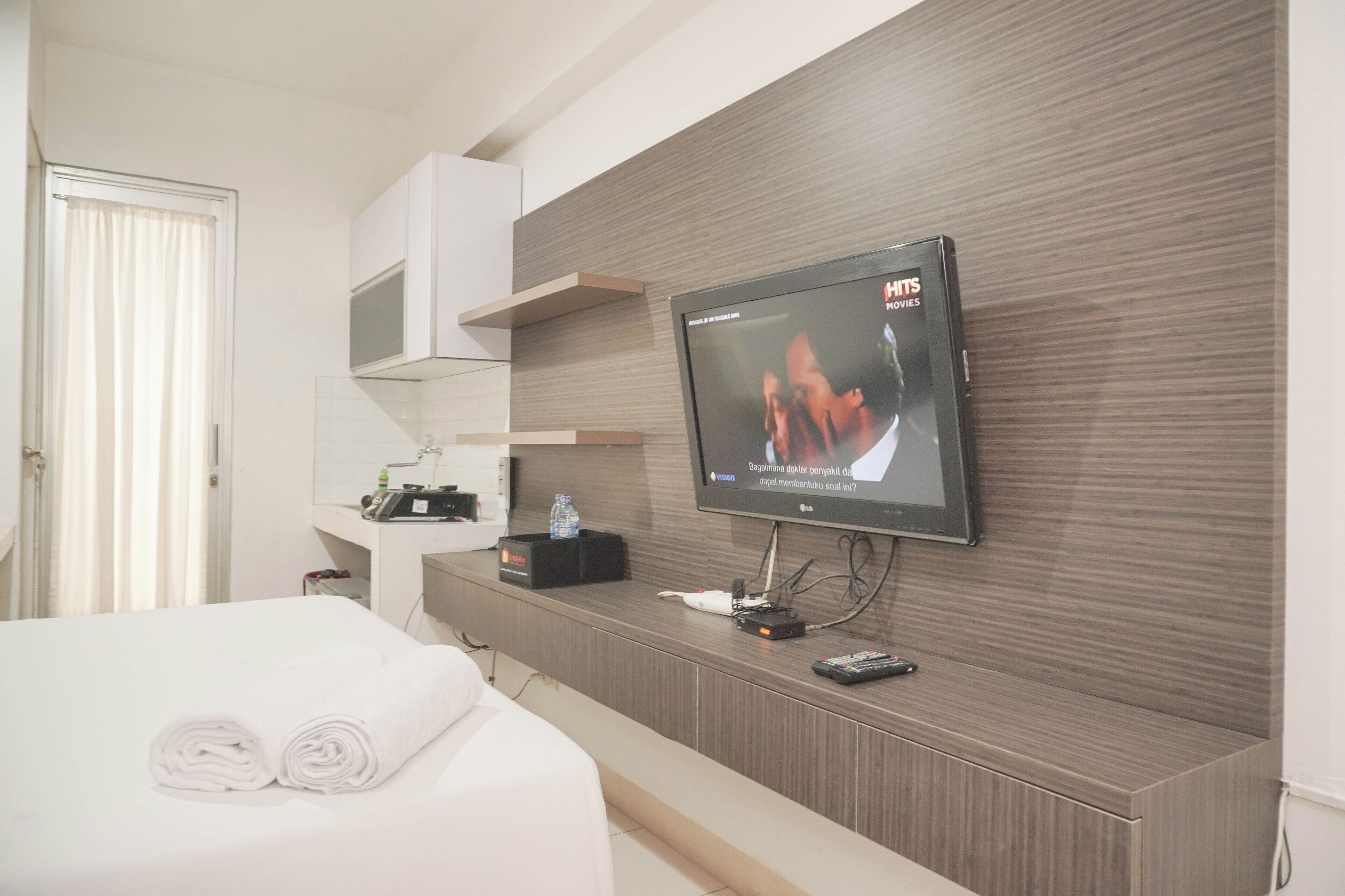 A hotel room with a flat-screen TV mounted on the wall, a bed with neatly folded white towels, a bedside table with a bottle and a remote control, and a wooden panel wall.